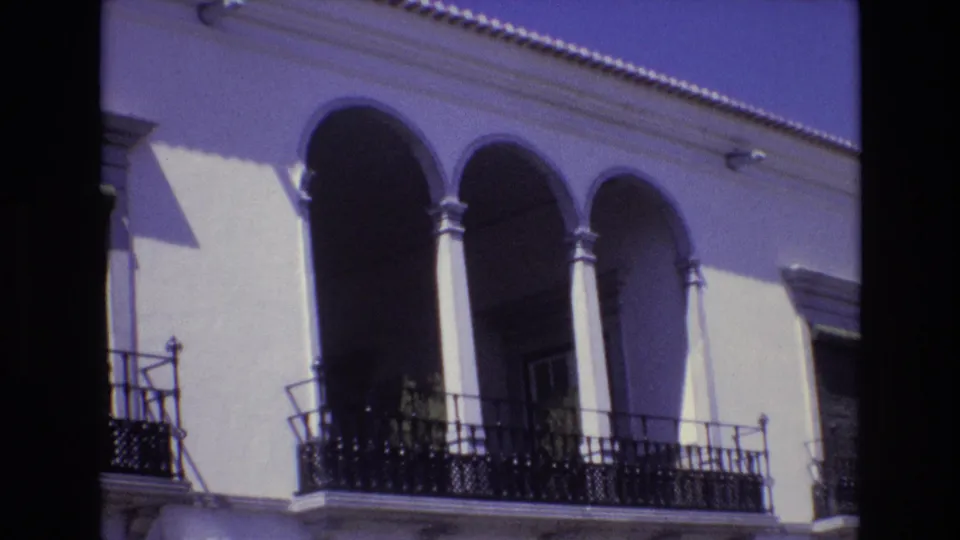 1972 Portugal 8mm frame showing a slightly shifted view of arched columns, black balcony railings, a pale wall, and strong shadows under the arches.