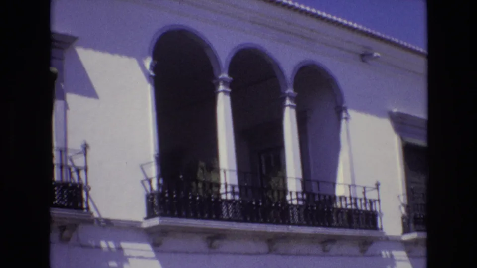1972 Portugal home-movie frame of the same arched building facade, with wrought-iron balcony railings, column shadows, roofline tiles, and a purple-blue film cast.
