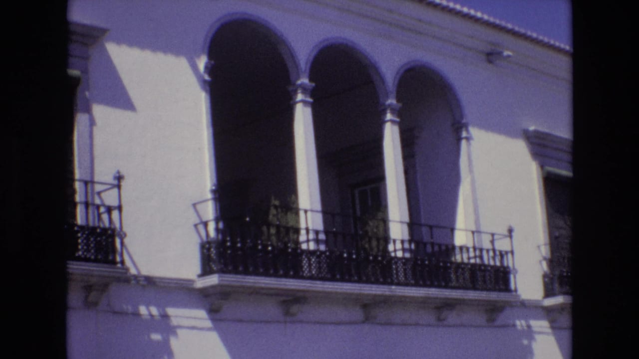 Portugal 1972 home-movie frame showing a sunlit white facade with arched balcony openings, slim columns, and ornate black iron railings.