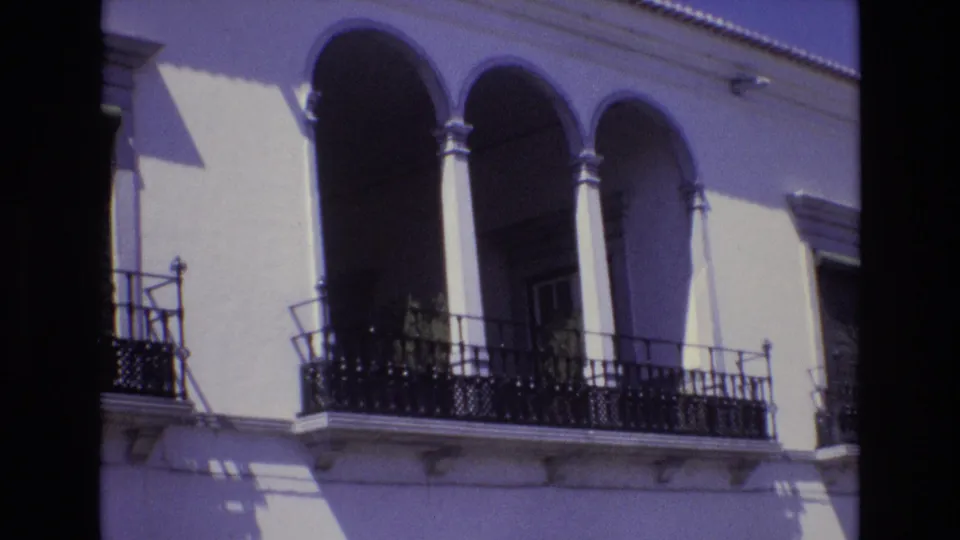 Portugal 1972 home-movie frame showing a sunlit white facade with arched balcony openings, slim columns, and ornate black iron railings.