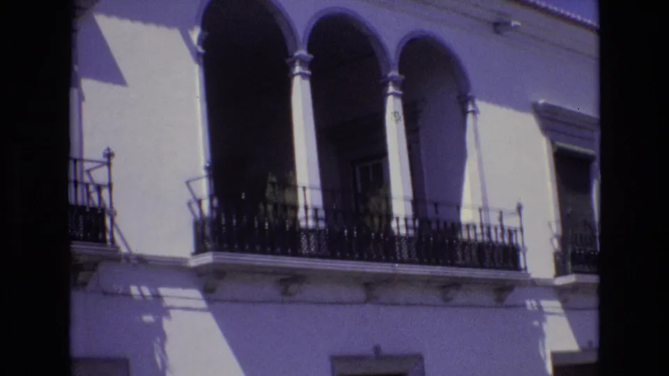 Portugal 1972 frame with a closer view of a white building balcony, arched openings, black ironwork, and hard afternoon-looking shadows.