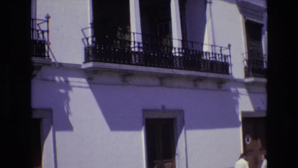 Portugal 1972 home-movie frame of the white balcony facade, with black iron railings, arched openings, a lower doorway, and a partially visible person near the bottom right edge.