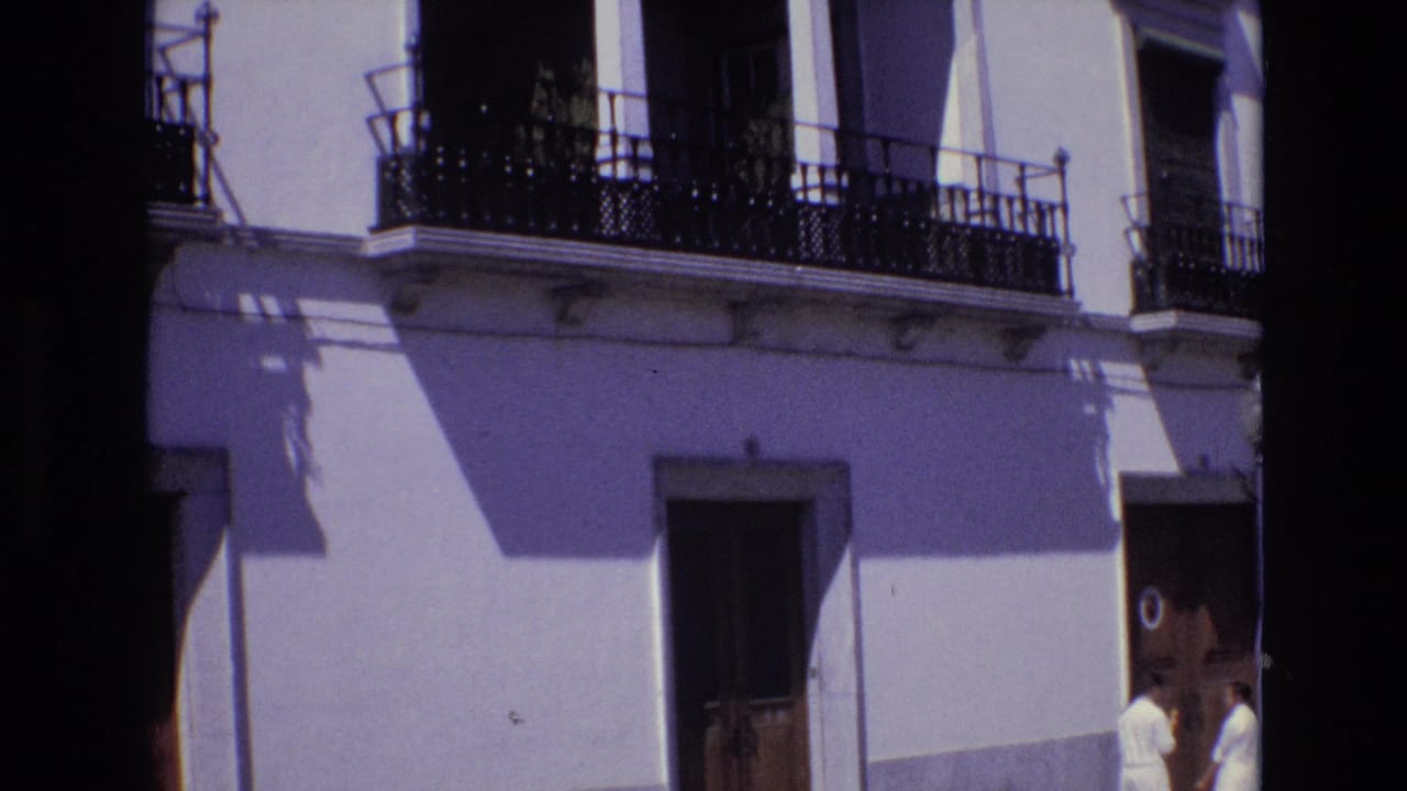 1972 Portugal street view showing a white stucco facade with black wrought-iron balconies, wooden doors, and two women in white dresses near the sidewalk.