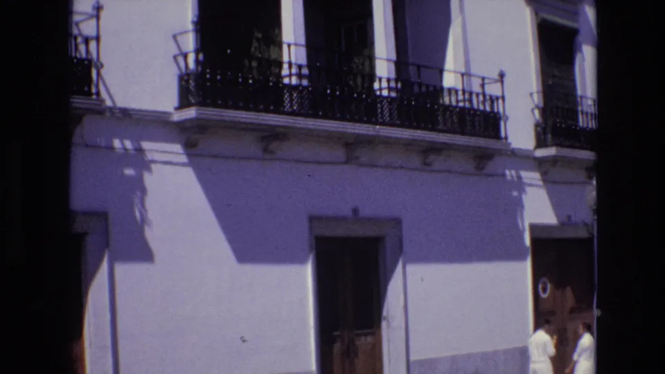1972 Portugal street view showing a white stucco facade with black wrought-iron balconies, wooden doors, and two women in white dresses near the sidewalk.