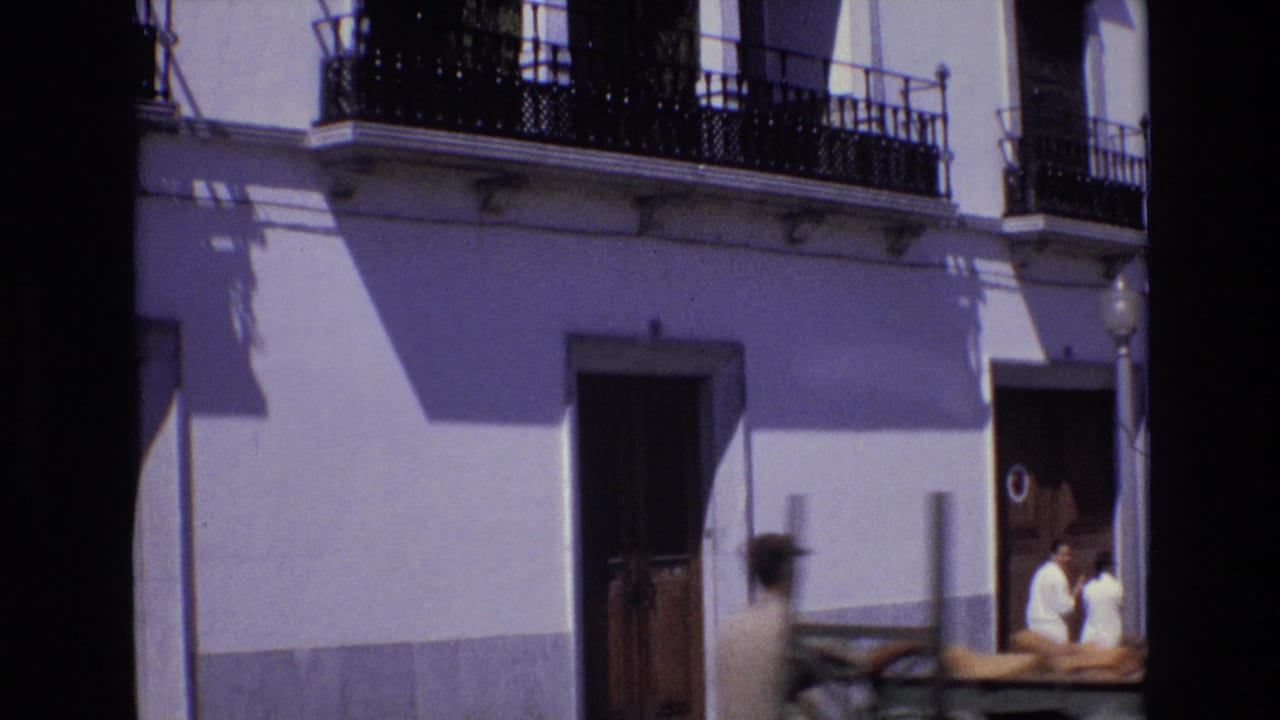 1972 Portugal street frame with a white stucco building, wrought-iron balconies, two pedestrians at the right edge, and a blurred passing vehicle.