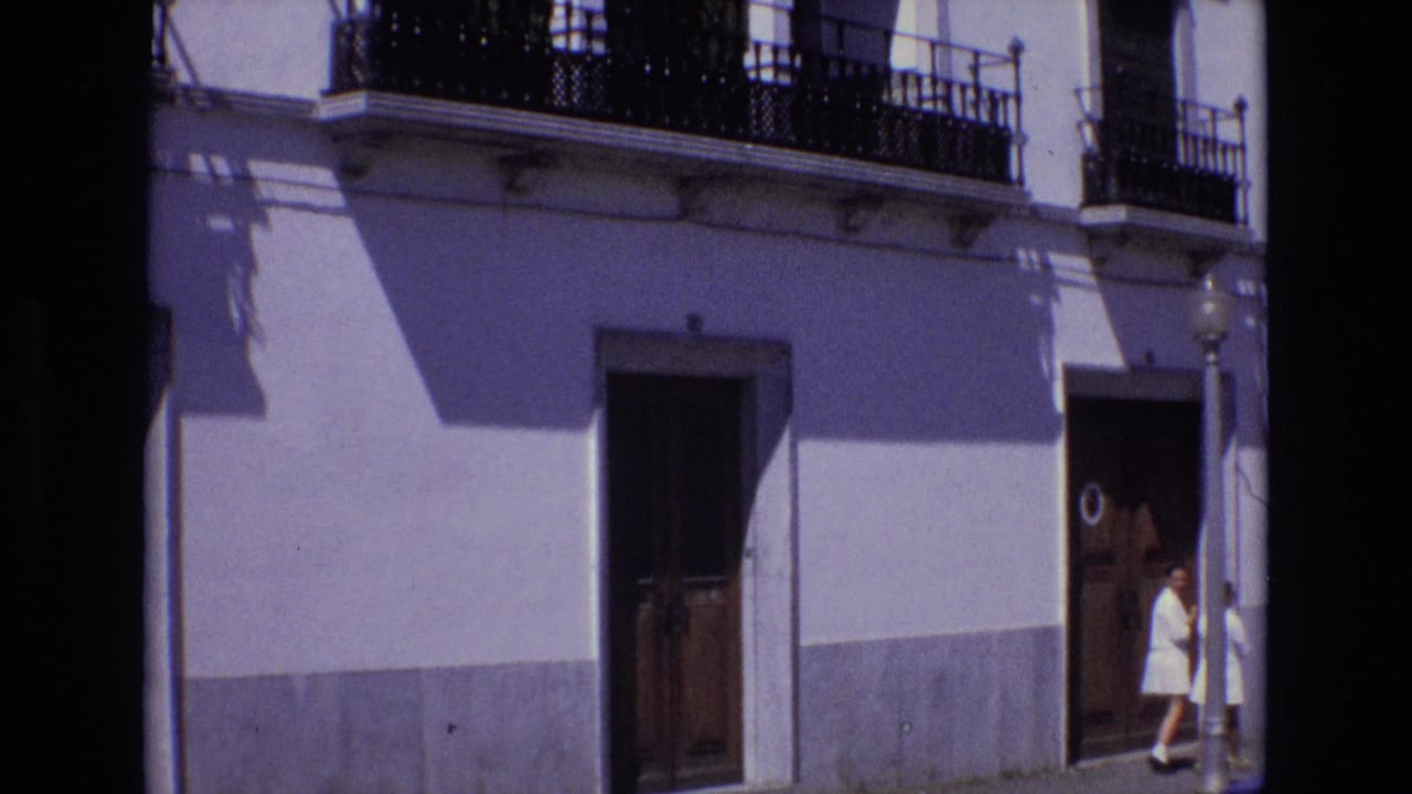 Two women in white dresses walk beside a white building with balconies and a street lamp.