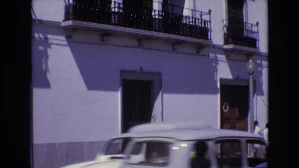1972 Portugal street frame showing a white vintage car moving across the foreground in front of a stucco building with balconies and pedestrians near a doorway.