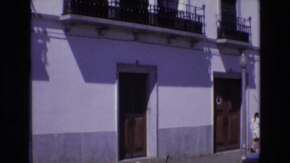 1972 Portugal 8mm frame showing a sunlit whitewashed street facade with black wrought-iron balconies, tall wooden doors, a streetlamp, and a person partly visible near the right edge.