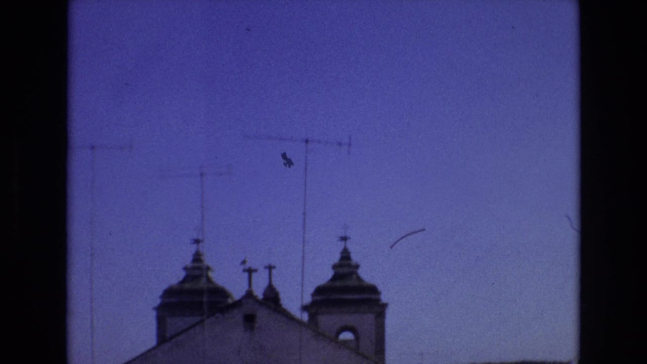 1972 Portugal 8mm frame showing a church roofline with twin domed towers and cross-topped details below a deep blue sky crossed by rooftop television antennas.