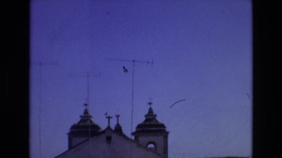 1972 Portugal 8mm frame showing a church roofline with twin domed towers and cross-topped details below a deep blue sky crossed by rooftop television antennas.