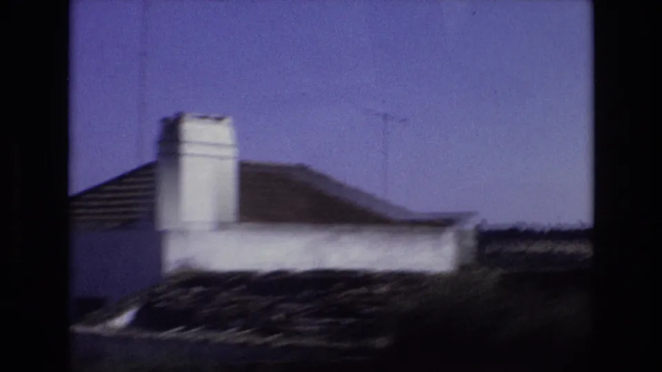 1972 Portugal 8mm frame showing a low white building or rooftop with a prominent chimney, dark tiled roofline, and blue sky in a soft handheld view.