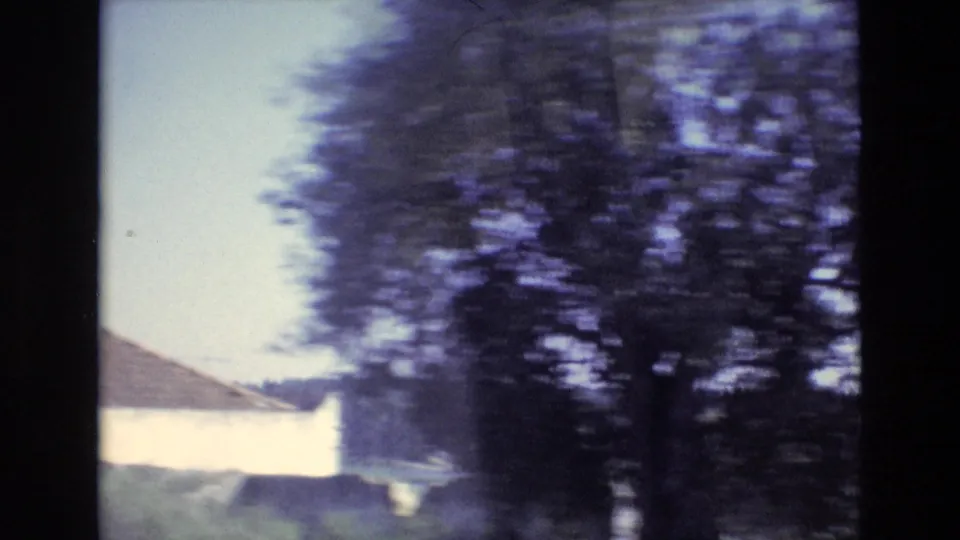 1972 Portugal home-movie frame showing blurred roadside trees, a pale sky, and part of a white rural building viewed from a moving vehicle.