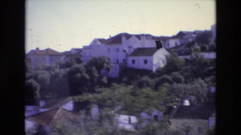 1972 Portugal travel frame showing whitewashed houses, dense greenery, tiled roofs, and a sloped residential area from a moving viewpoint.