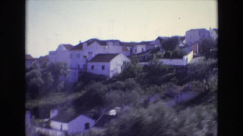 1972 Portugal home-movie frame with hillside homes, trees, rooftop antennas, and blurred foreground rooftops from a train-window view.