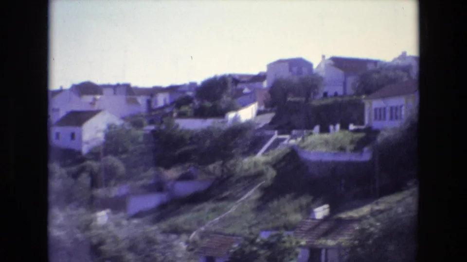 1972 Portugal travel footage frame with white houses, stepped garden terraces, trees, and a sunlit hillside viewed across the valley.