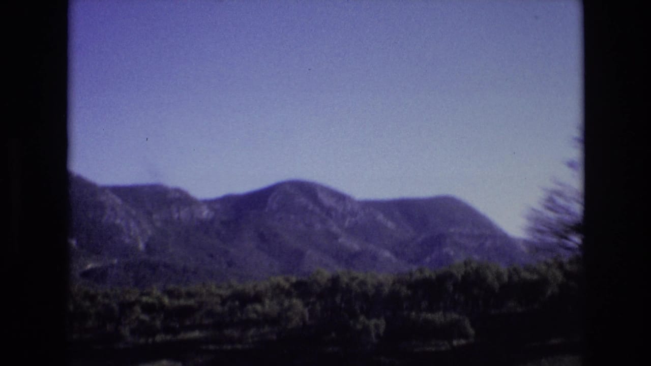 1972 Portugal travel footage frame with a distant mountain range beyond dark green wooded terrain, seen from a moving vehicle.