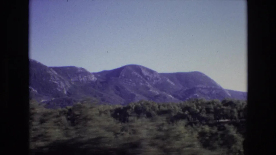 1972 Portugal frame showing a broad mountain ridge behind wooded slopes and roadside greenery, with the moving-vehicle viewpoint still apparent.