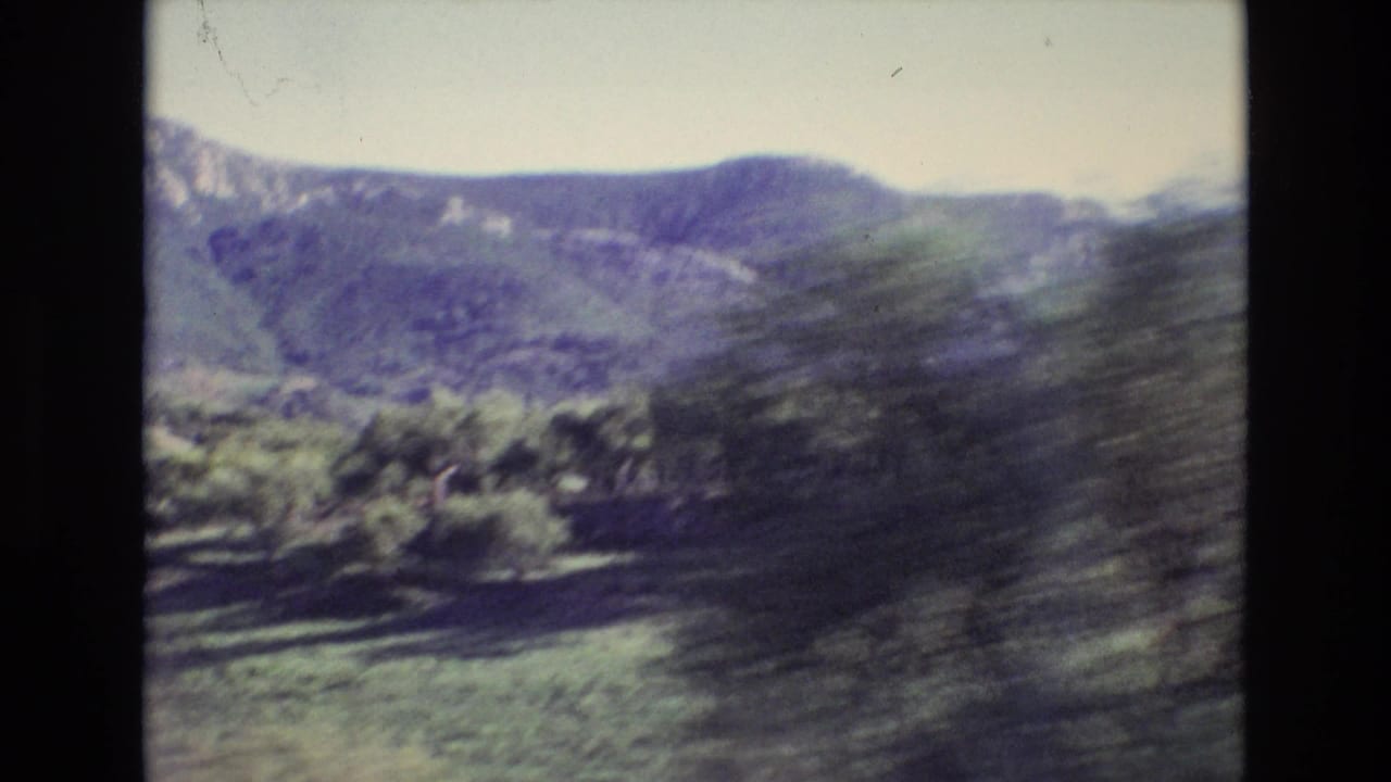 Portugal 1972 8mm travel frame showing a distant green mountain ridge and pale sky, with roadside foliage blurred by vehicle movement.