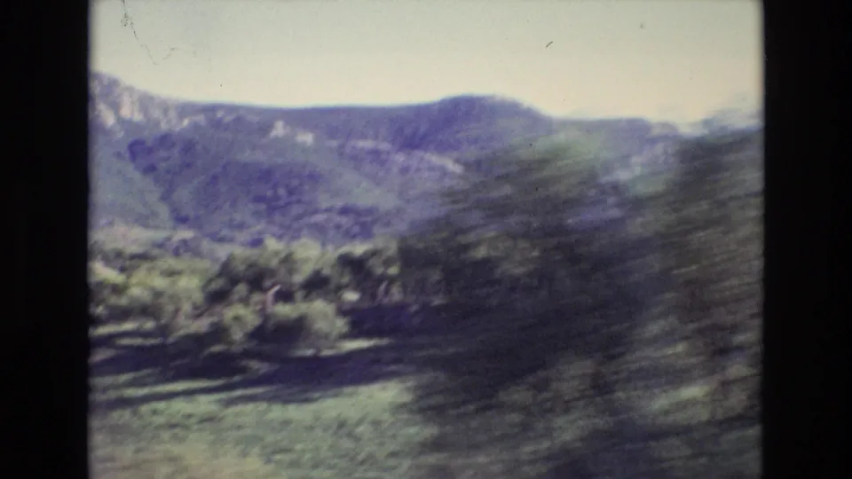 Portugal 1972 8mm travel frame showing a distant green mountain ridge and pale sky, with roadside foliage blurred by vehicle movement.