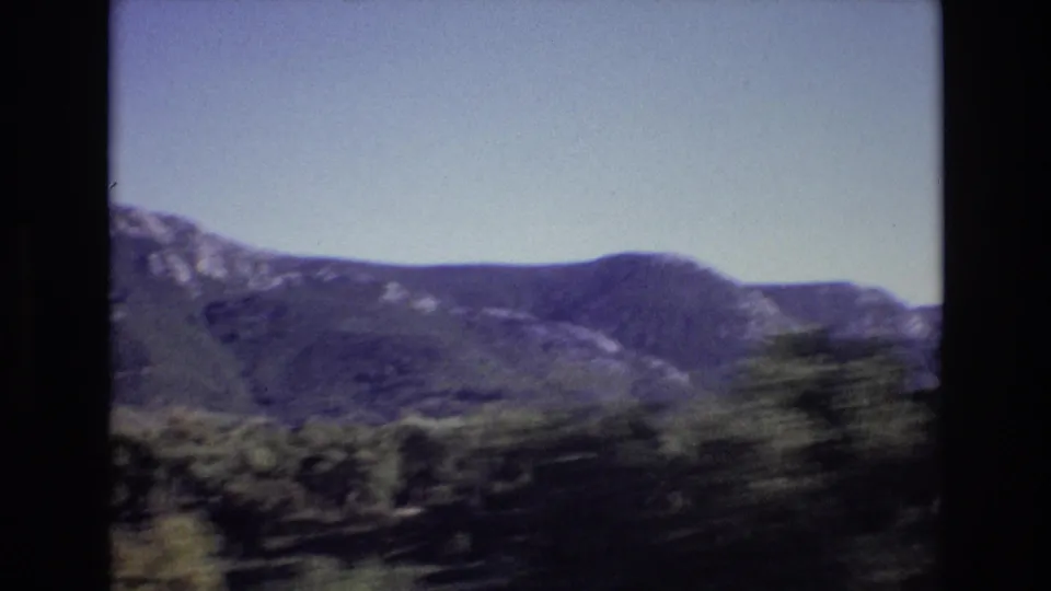 Portugal 1972 8mm frame looking across blurred roadside foliage toward distant rounded mountain ridges beneath a clear sky.