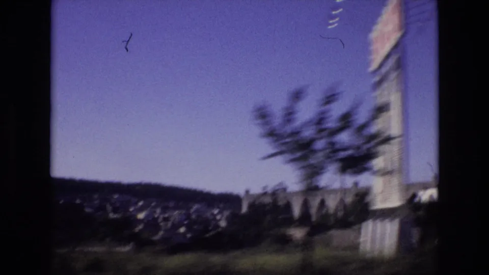 Portugal 1972 home-movie frame with a hillside settlement, distant ridge, blurred roadside tree, and the edge of a building passing the camera.
