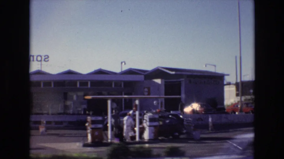Portugal 1972 home-movie frame showing a roadside fuel station, classic cars, and people standing near the pumps in strong sunlight.