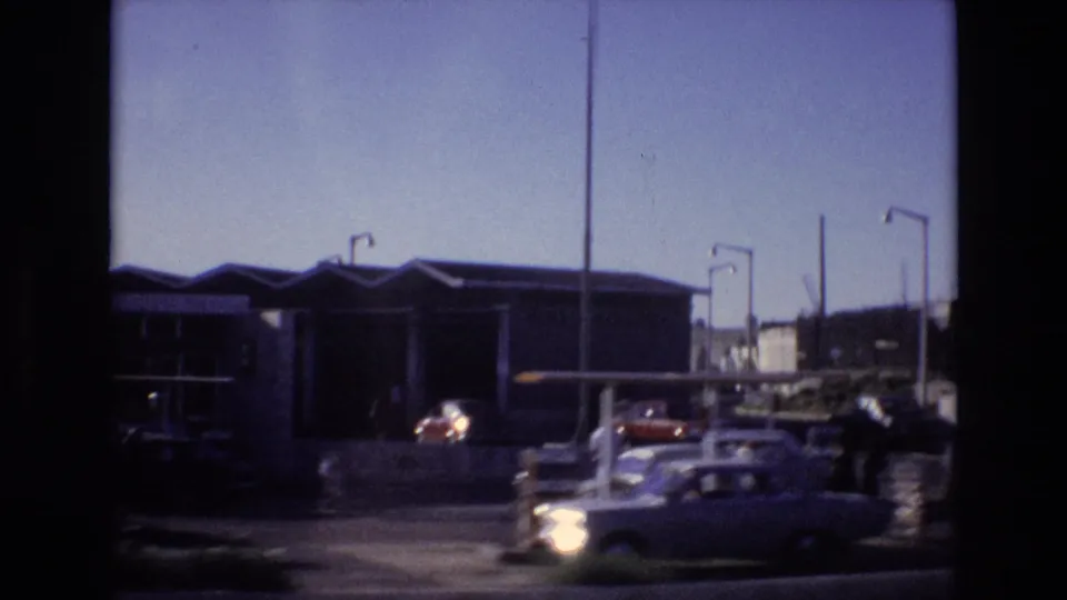 Portugal 1972 roadside scene with classic cars moving past a fuel station, people near the forecourt, and tall lamp posts against a pale sky.