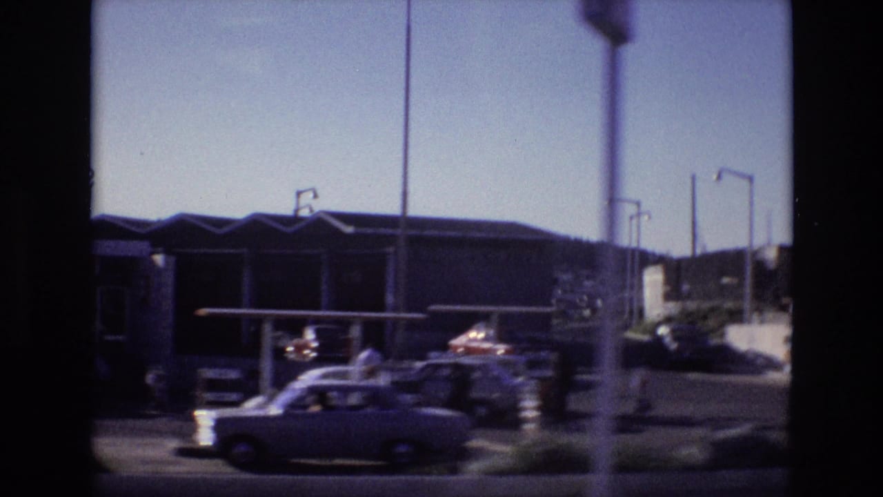 Parking area with small cars, streetlights, and a low building seen from a moving vehicle.