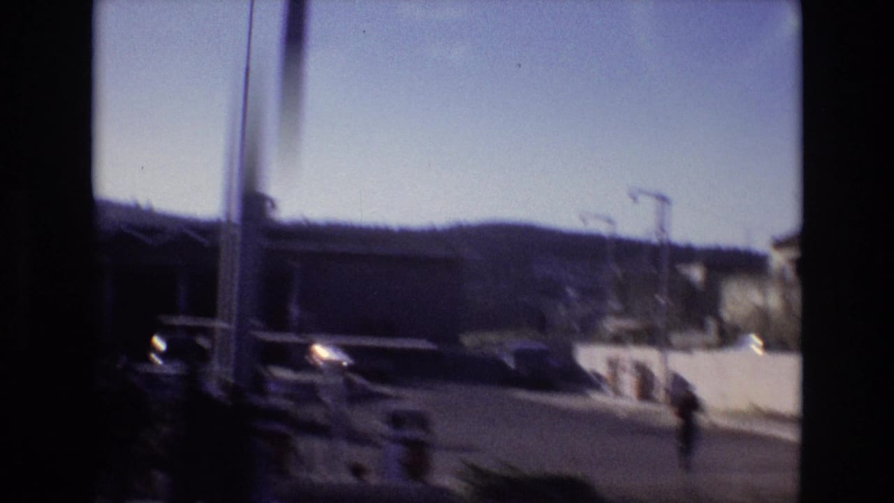 Roadside view with parked vehicles, streetlights, low buildings, and a pedestrian at the edge of frame.
