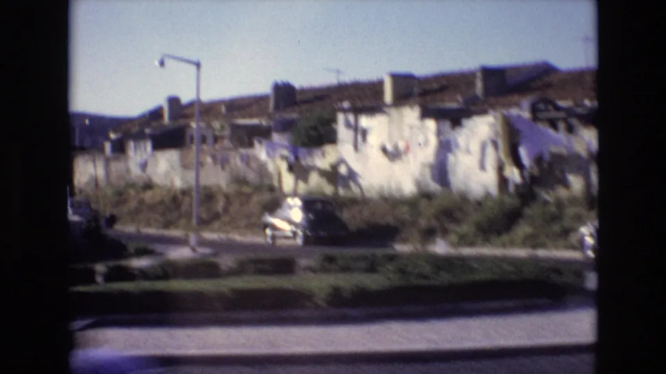 1972 Portugal residential roadside view with a long masonry wall, tiled roofs, hanging laundry, streetlights, low shrubs, and a parked car.