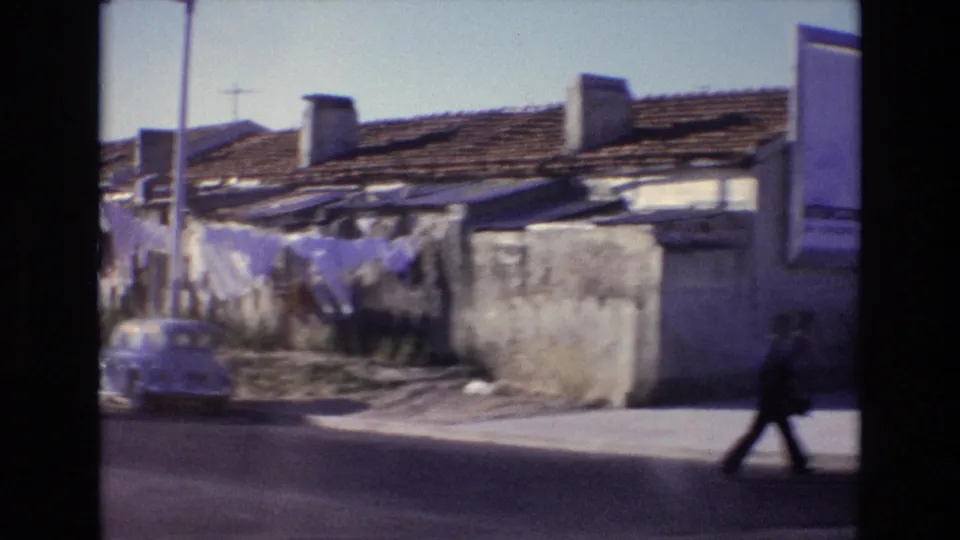 1972 Portugal urban residential street showing tiled roofs, masonry walls, a parked car, streetlight, and a distant pedestrian walking along the sidewalk.