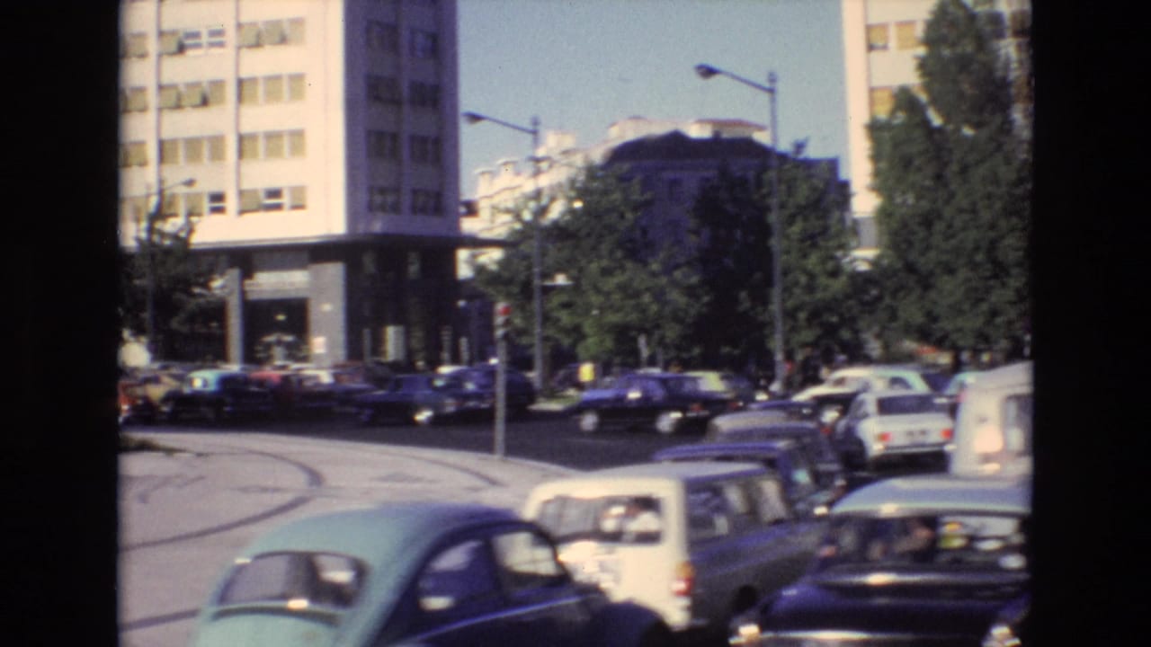 Portugal 1972 street view showing classic cars and a light-colored van moving through an urban intersection below a modern multi-story building.