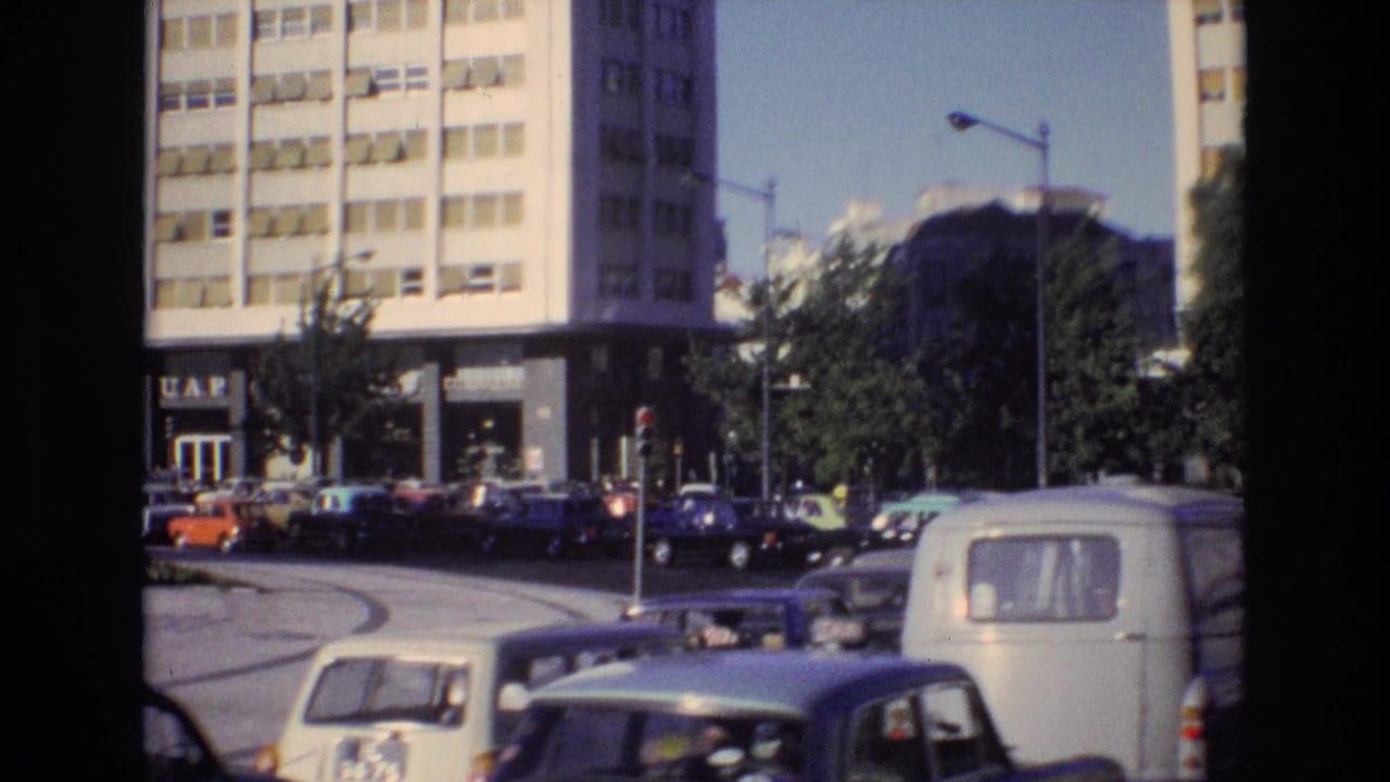 Portugal 1972 urban traffic scene with parked and moving cars near a building entrance bearing visible signage, with trees and streetlights nearby.