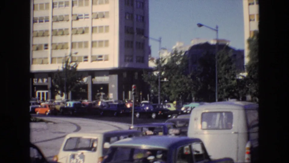 Portugal 1972 urban traffic scene with parked and moving cars near a building entrance bearing visible signage, with trees and streetlights nearby.