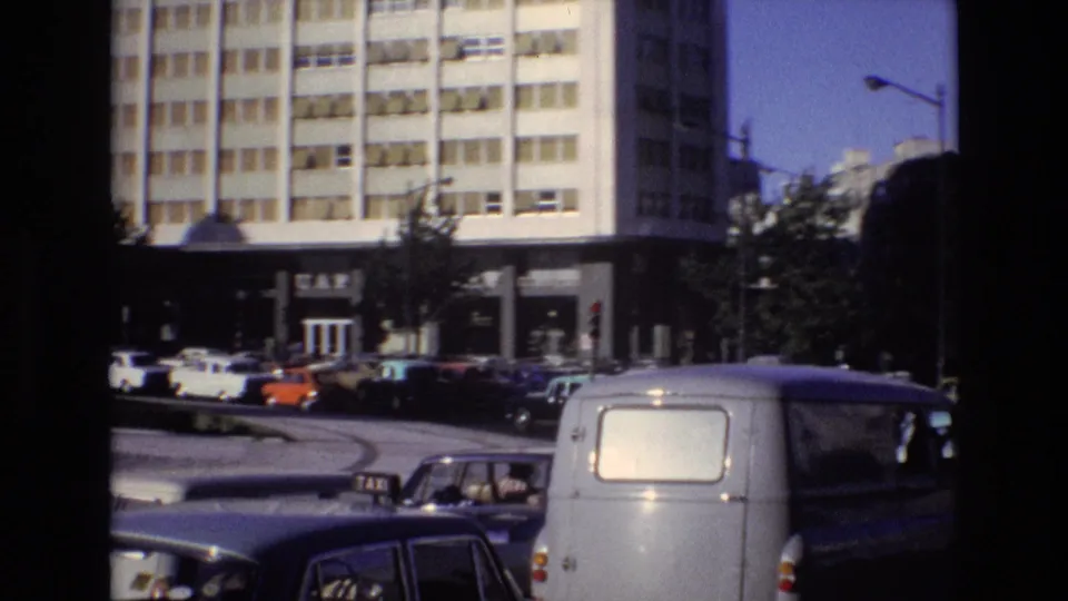 Portugal 1972 street frame showing a taxi-marked car, a light-colored van, several classic cars, and a modern grid-window building in the background.