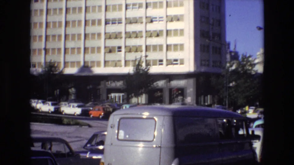 Portugal 1972 urban street view with a light-colored van in the foreground, classic cars nearby, and a tall modern building rising behind roadside trees.