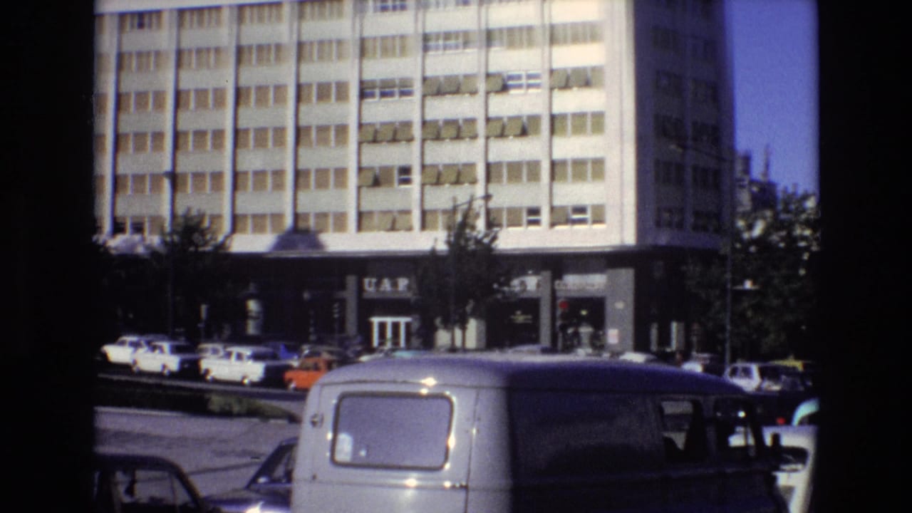 1972 Portugal 8mm frame showing a multi-story urban building with a visible UAP sign, parked cars, street trees, and a white van in the foreground.