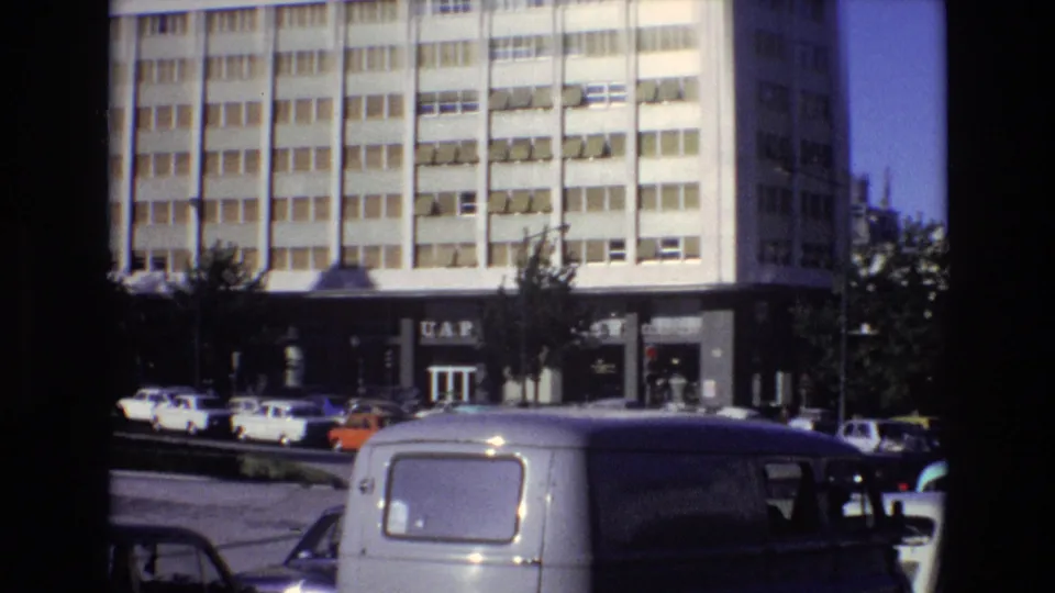 1972 Portugal 8mm frame showing a multi-story urban building with a visible UAP sign, parked cars, street trees, and a white van in the foreground.