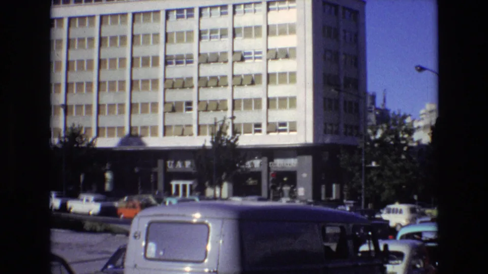 1972 Portugal street view with a large modernist building, visible signage, parked cars, and a white van partly blocking the lower foreground.
