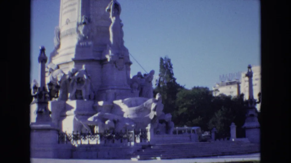 1972 Portugal visitor-shot frame of an ornate public stone monument with sculpted figures, steps, trees, and surrounding street elements under a clear sky.