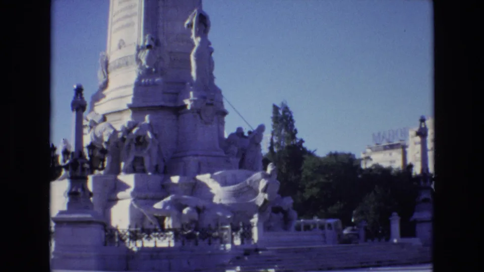 1972 Portugal 8mm frame showing a large sculptural monument with carved figures, railings, steps, trees, and a small vehicle near the street edge.