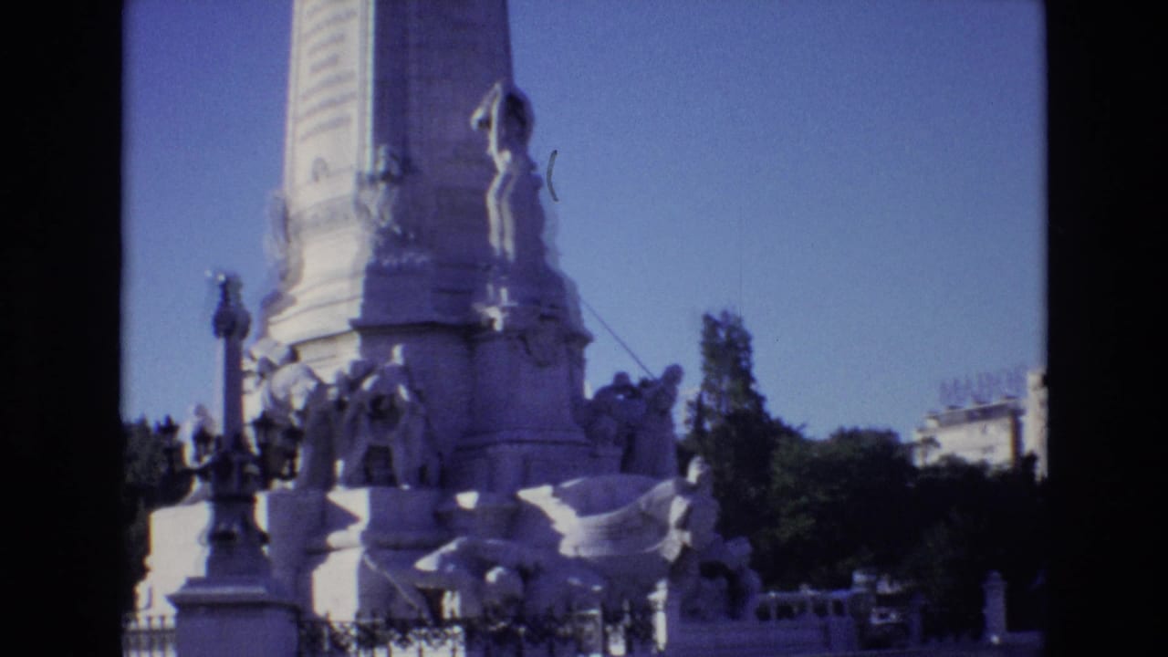 1972 Portugal 8mm frame showing the lower section of a pale stone obelisk with classical sculpted figures at its base under a clear blue sky.