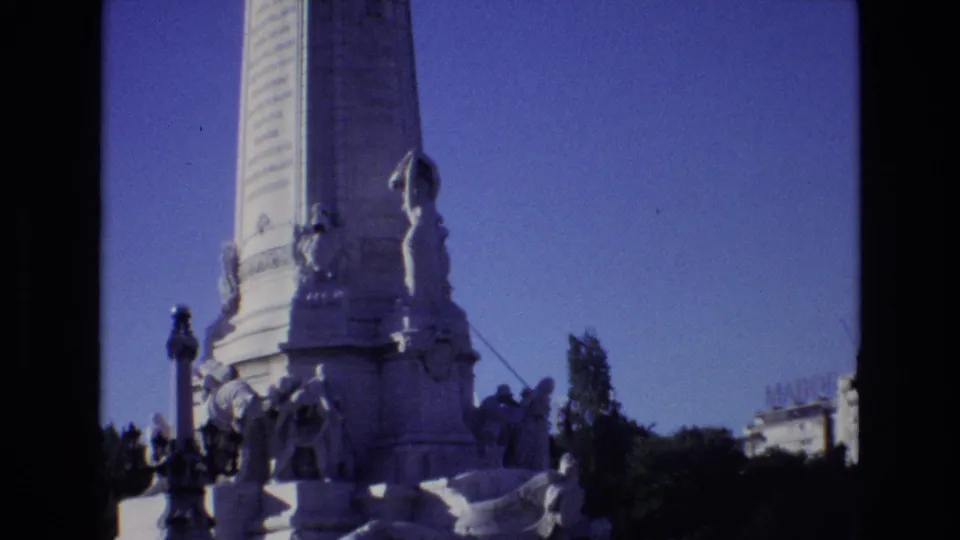 1972 Portugal home-movie frame of a tall stone monument rising from a sculpted base, with trees and a nearby building edge in the background.