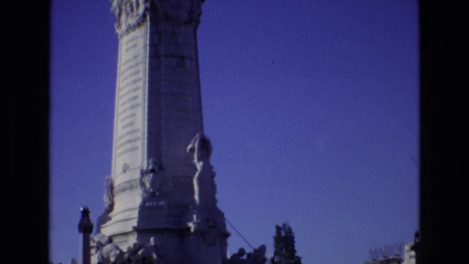 Final 1972 Portugal home-movie frame showing the tall obelisk and sculpted figures near its base, framed by a clear blue sky and dark 8mm borders.