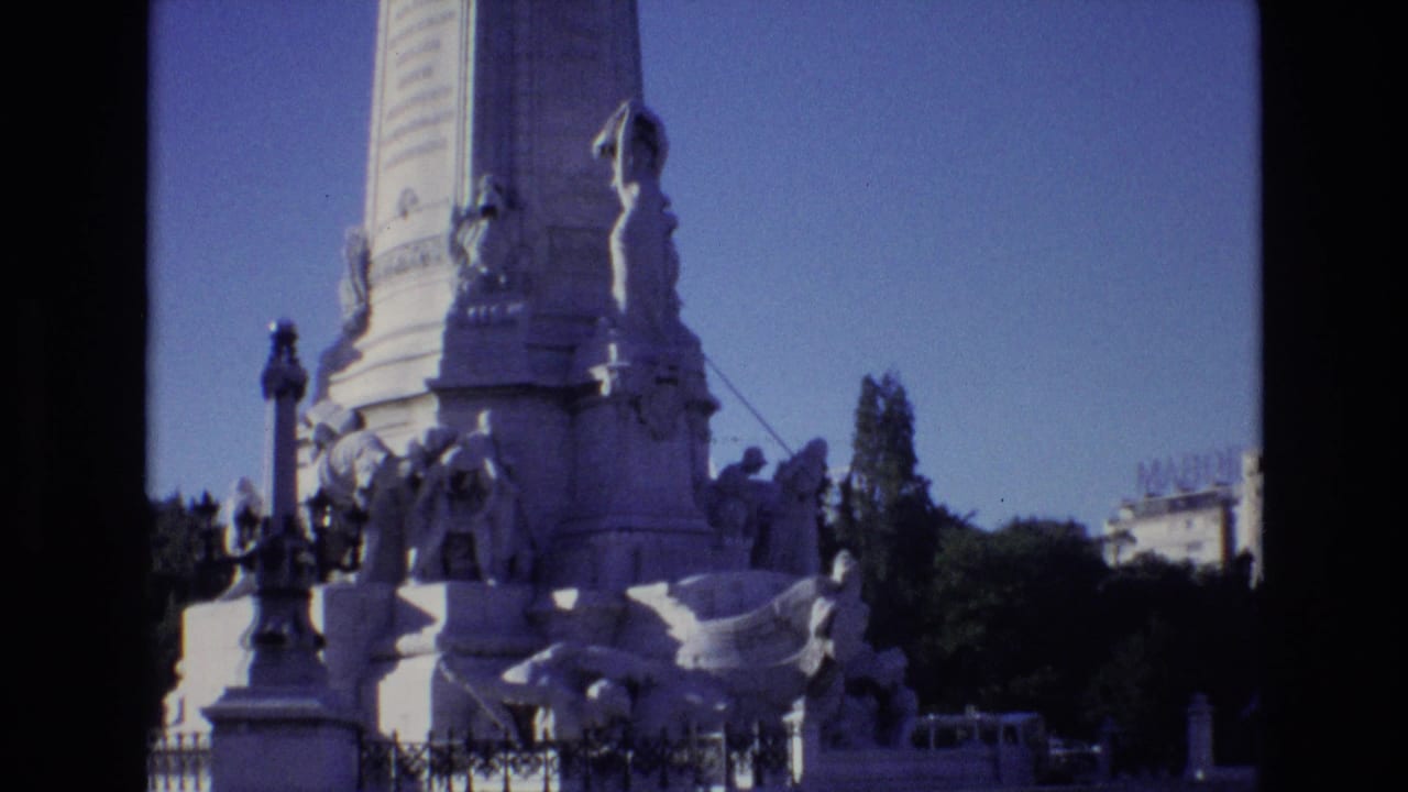 Ornate monument base with carved figures, lamps, trees, and distant buildings under a clear sky.