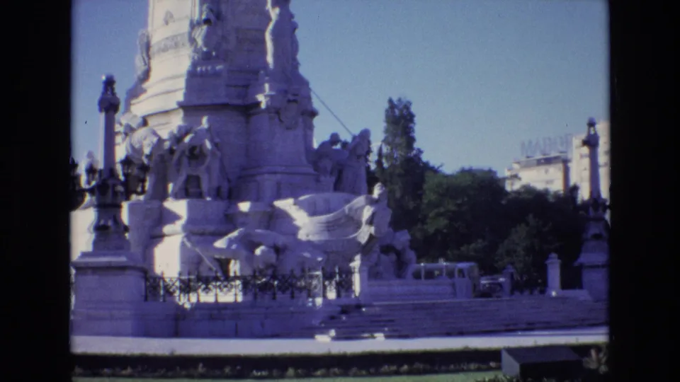 1972 Portugal 8mm frame showing a wider visitor-shot view of the stone monument base, sculpted figures, iron fencing, trees, and distant rooftop lettering.