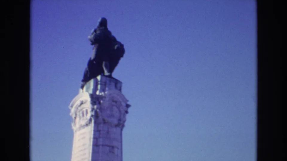 1972 Portugal 8mm frame isolating the upper section of the tall stone monument, with carved ornament and a dark statue silhouetted against the sky.