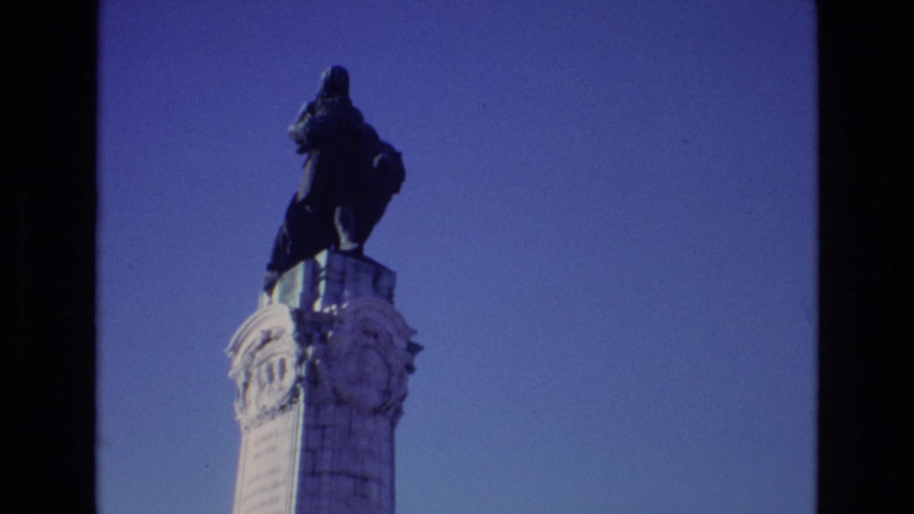 1972 Portugal 8mm frame showing a dark statue on a carved white pillar, filmed from below against a clear blue sky.