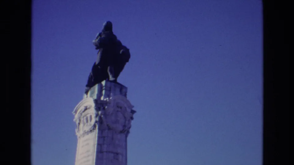 1972 Portugal 8mm frame showing a dark statue on a carved white pillar, filmed from below against a clear blue sky.
