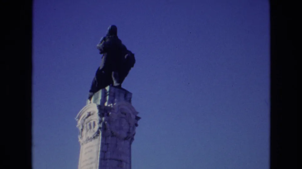 1972 Portugal home-movie frame with a dark monument figure above an ornate white column, seen in soft 8mm color.